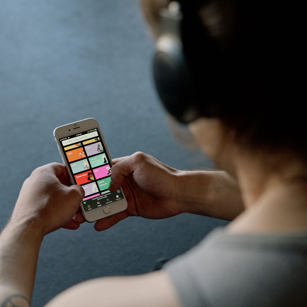 A person wearing headphones is focused on selecting music from a colorful app interface on a smartphone, the screen displaying various playlist options. The hands show light tattoos, suggesting a casual or creative lifestyle.