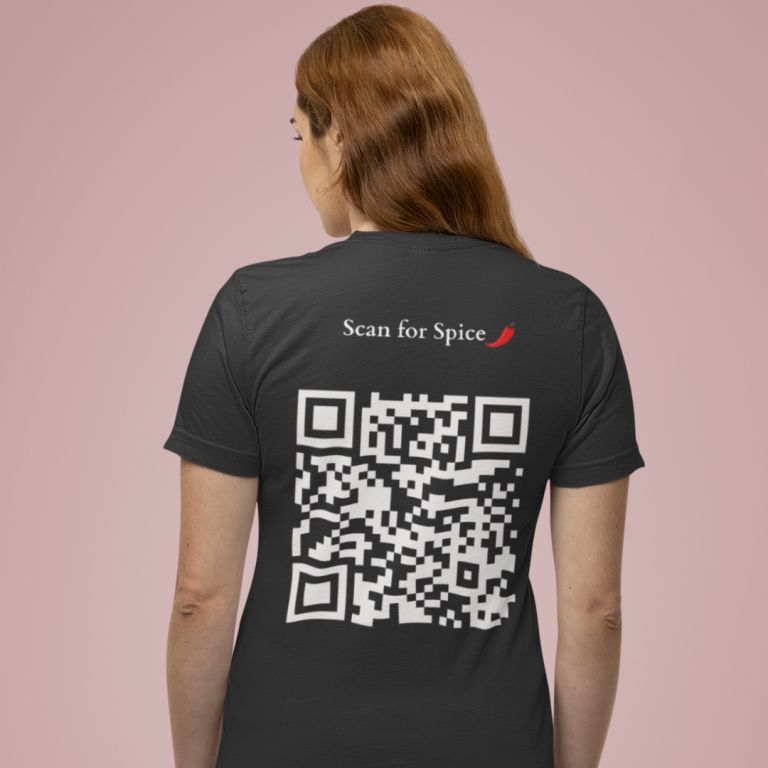 A woman seen from behind wears a black t-shirt with "Scan for Spice" and a large QR code printed on the upper back, accessorized with a red chili graphic, standing against a soft pink background.