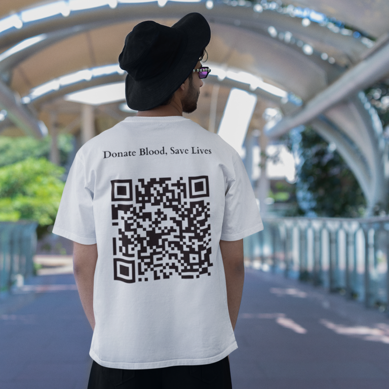 A man stands with his back to the camera, wearing a white t-shirt with "Donate Blood, Save Lives" above a QR code, a black bucket hat, black pants, against an architectural backdrop with an overhead canopy.