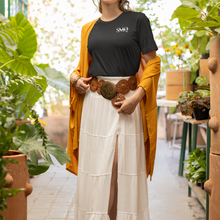 A woman stylishly dressed in a black SMQ logo t-shirt paired with a long white skirt and a mustard yellow cardigan, posing in a lush greenhouse setting.