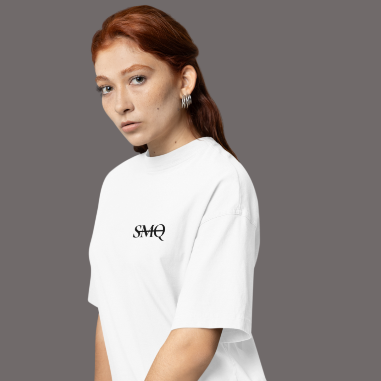 A young woman with red hair and a subtle gaze, modeling a white t-shirt adorned with the black lettered monogram 'SMQ' on the chest.