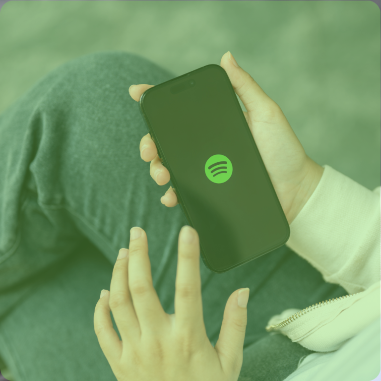 A close-up of a person's hands holding a smartphone displaying the Spotify logo, set against a green-toned background that matches their casual outfit.