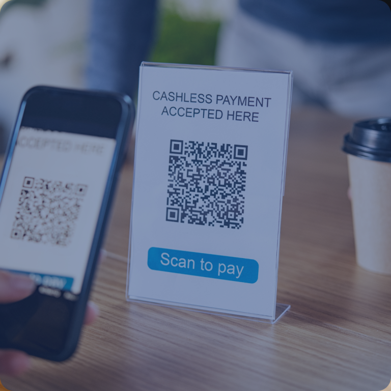 A hand holds a smartphone scanning a QR code labeled "Cashless Payment Accepted Here" on a table placard, with a coffee cup nearby, suggesting a payment process at a cafe.